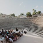 Bride and groom exchanging vows during a wedding ceremony at the Altos de Chavón Roman-style amphitheater at Casa de Campo in the Dominican Republic, with guests seated on stone steps at sunset.