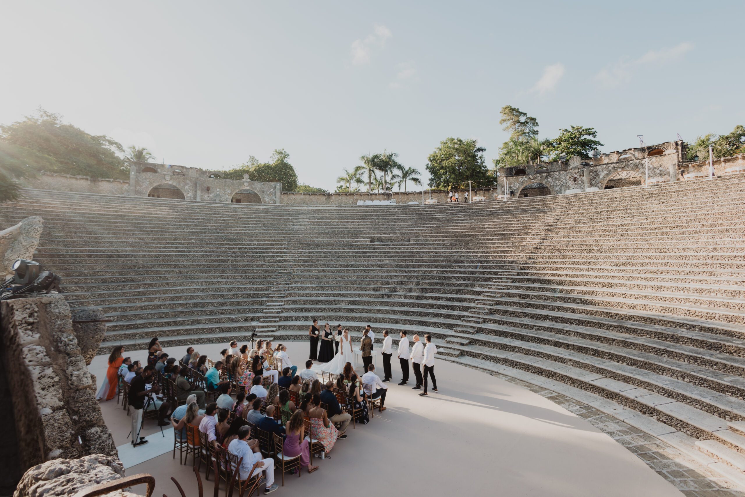 Bride and groom exchanging vows during a wedding ceremony at the Altos de Chavón Roman-style amphitheater at Casa de Campo in the Dominican Republic, with guests seated on stone steps at sunset.